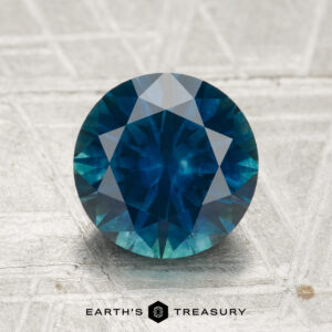A 2.32-carat heated Montana sapphire, round and brilliant-cut, rests on a textured metallic surface with the "EARTH'S TREASURY" logo and name at the bottom.
