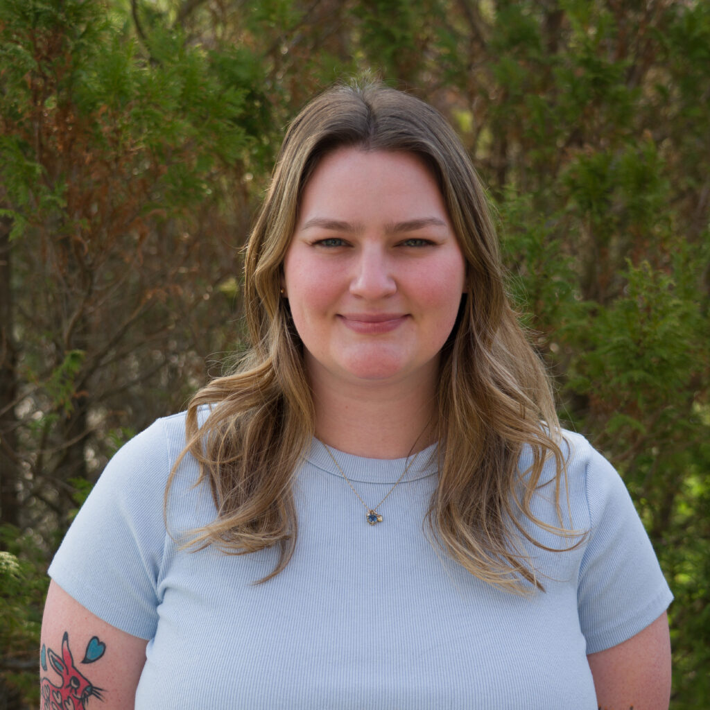 A woman with long, light brown hair smiles at the camera outdoors, wearing a light blue short-sleeve shirt in front of green foliage. Product: _DSC0237.