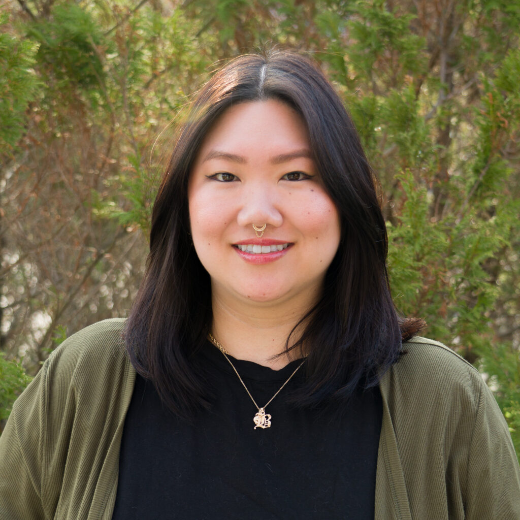 Wearing a septum ring, necklace, black shirt, and green jacket, a person with long dark hair smiles at the camera outdoors in front of greenery in photo _DSC0275.