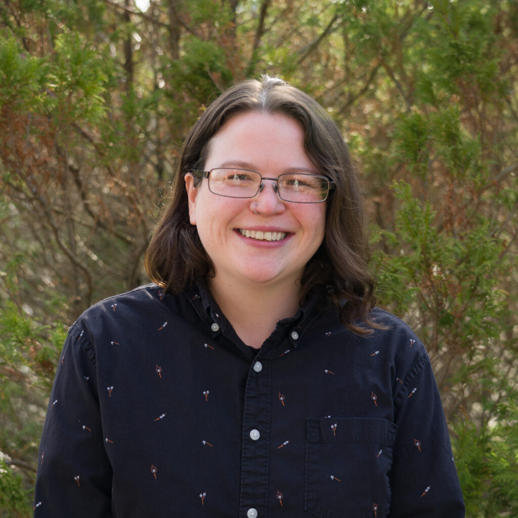 A person with glasses and shoulder-length brown hair smiles outdoors before green foliage, wearing the _DSC0318 dark button-up shirt featuring a small pattern.