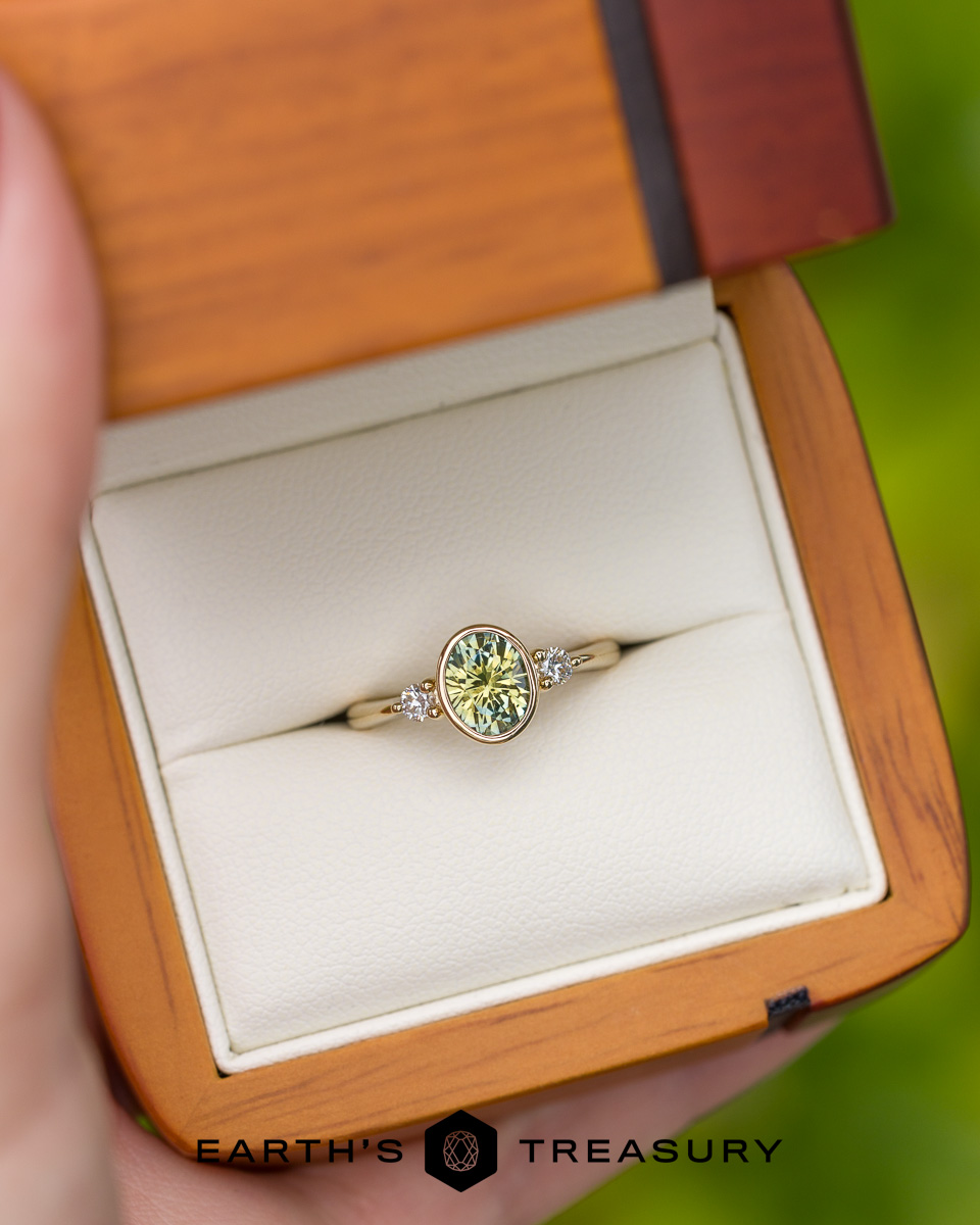The _DSC3763-1200-BW gold ring features an oval green gemstone and side diamonds, showcased in a wooden jewelry box with white padding. "Earth's Treasury" is printed below, offering a stunning choice inspired by our Engagement Ring Guide.