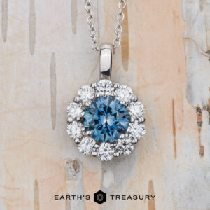 The "Dazzle" necklace in 14K white gold with a 6.0mm medium blue Montana sapphire center and a halo of clear stones is shown on a light wood background, with the Earth’s Treasury logo at the bottom.