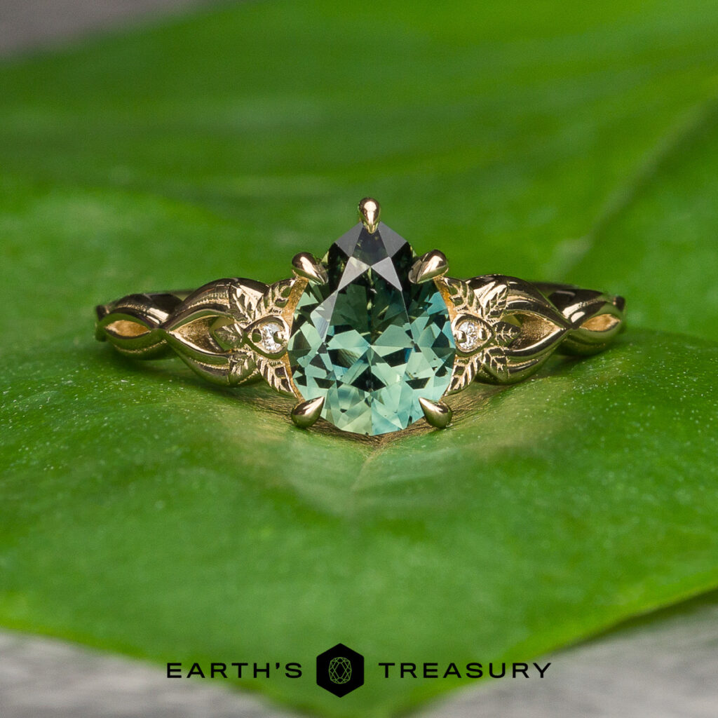 A pear-shaped green gemstone ring from the Williams, George [custom gallery], featuring gold and diamond accents, rests on a green leaf with the Earth’s Treasury logo below.