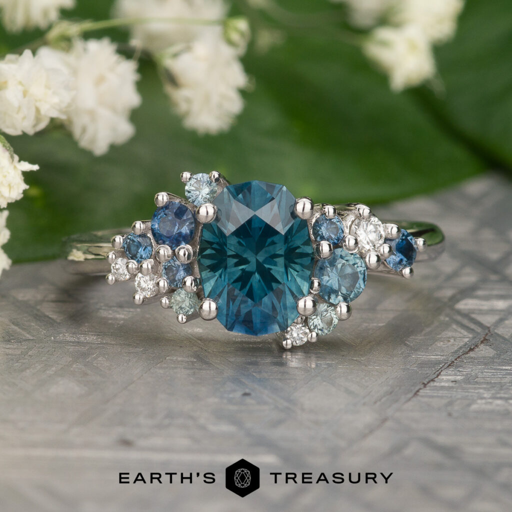 The Figgess, Liana ring showcases a large blue oval gemstone set in silver, accented by smaller blue and white stones, and is presented on a textured surface with flowers and green leaves in the background.