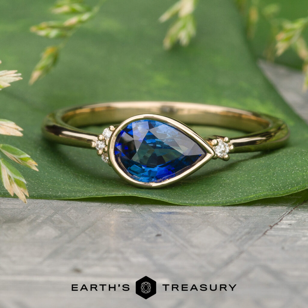 Gold ring featuring a pear-shaped blue gemstone center and small accent stones, displayed on green with plant details; "Earth's Treasury" logo beneath. Product: Ortiz, Maria [custom gallery].