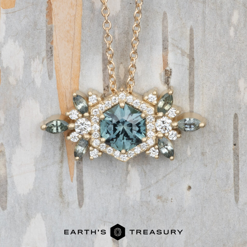 The West, Victoria 3 necklace features a hexagonal teal gemstone pendant accented by white and marquise-shaped teal stones on a gold chain, shown on a wood background. Earth's Treasury logo appears below.