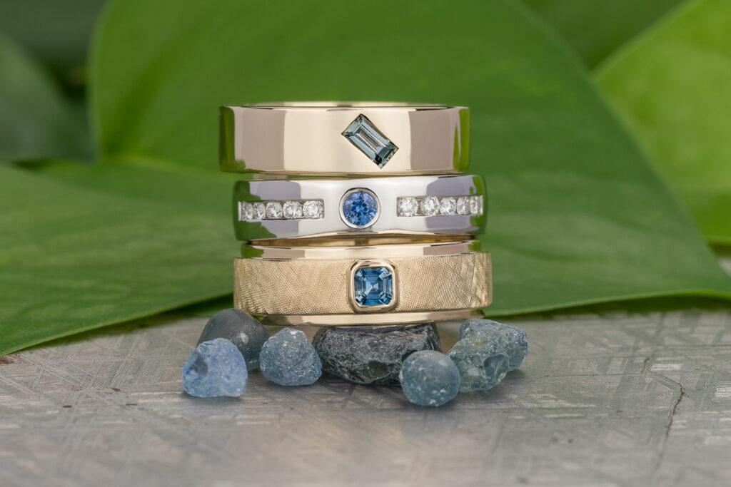 The _SNY1220-Edit-3-1200-BW features three gold and silver rings set with blue gemstones and diamonds, stacked on blue stones with green leaves in the background.