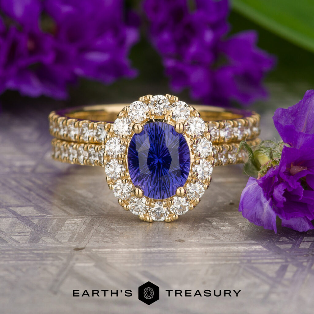 The Dworak, Alexander (Gustav) 1 ring features a blue gemstone center encircled by diamonds. It rests on a patterned surface adorned with purple flowers, with the "Earth's Treasury" logo displayed at the bottom.