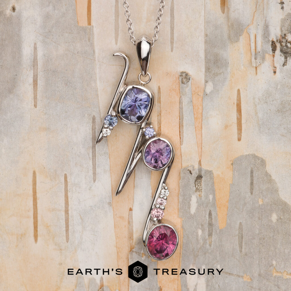 A silver pendant necklace with three round blue, purple, and pink gemstones, accented by clear stones, displayed on light wood. Text reads "McCue, Megan [custom library].