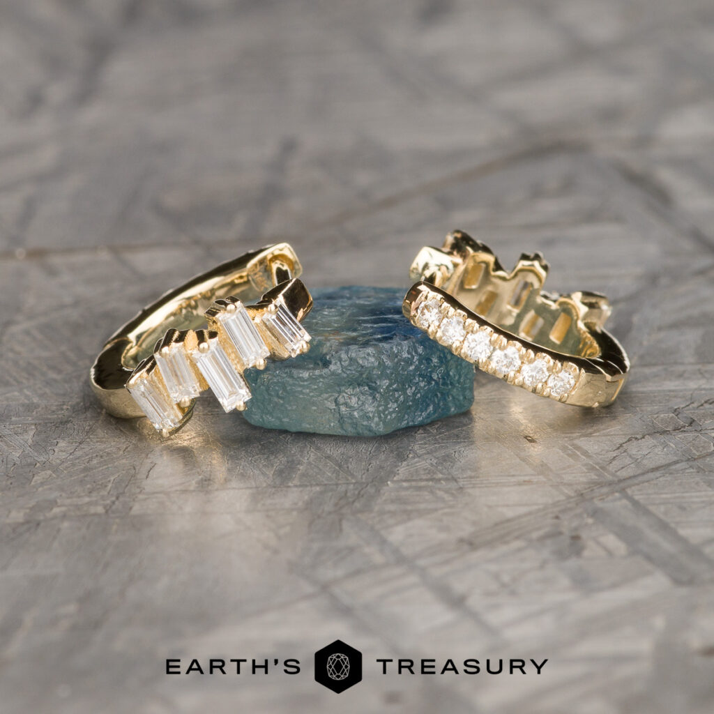Two gold rings with gemstones from the "Mielke, Christine 5 [custom gallery]" collection are shown leaning against a rough blue stone on a textured gray surface, with the “Earth’s Treasury” logo at the bottom.