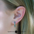 Close-up of a person's ear wearing eclipse hoops, featuring five blue-green gemstones; the "Earth's Treasury" logo appears at the image's bottom.