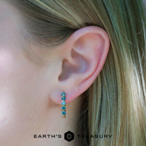 Close-up of a person's ear wearing eclipse hoops, featuring five blue-green gemstones; the "Earth's Treasury" logo appears at the image's bottom.