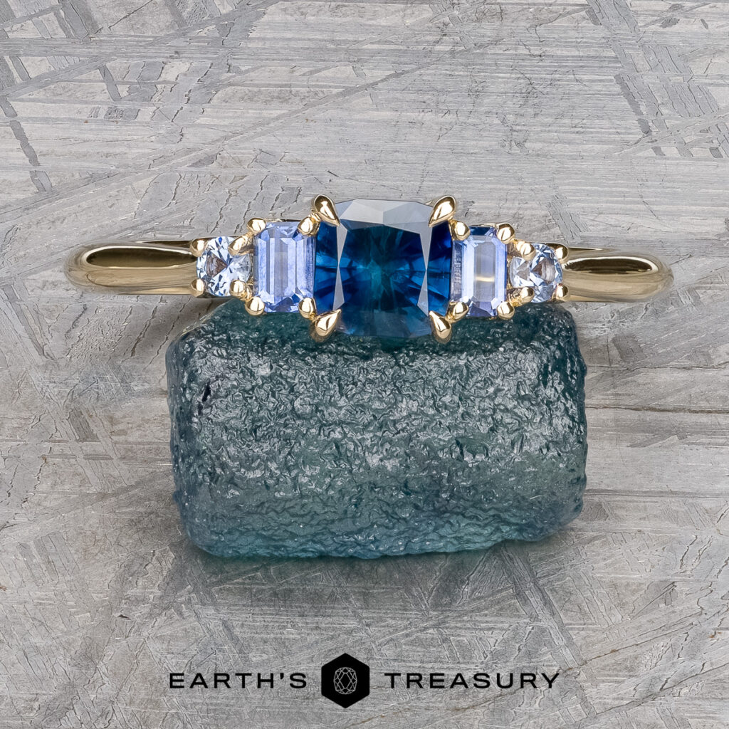 Noel, Nakita and Mitchell, Denise 1 [custom gallery]: A gold ring featuring a blue center gemstone and clear side stones is displayed on a rough blue crystal, with the "Earth's Treasury" logo beneath.