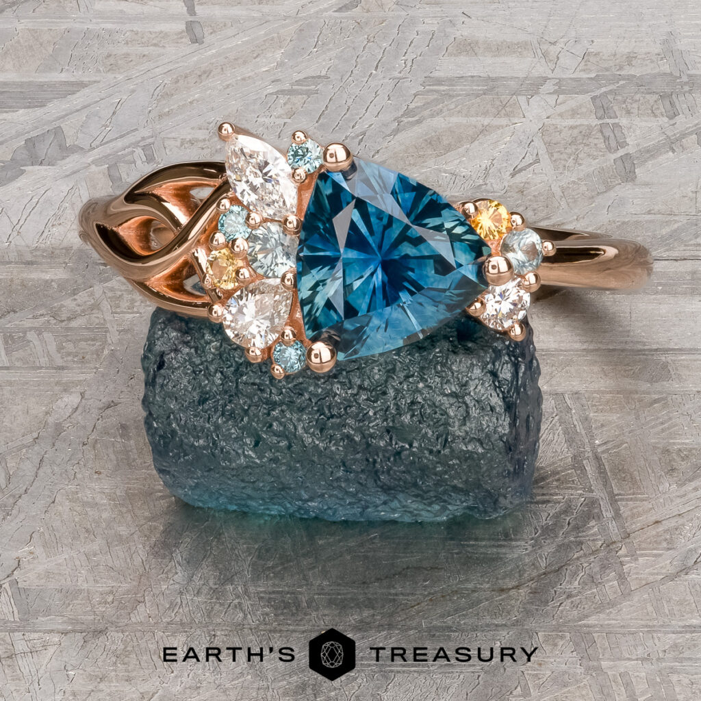 The Kazakoff, Miroslav (Miro) and Lee, Renee [custom gallery] features a rose gold ring with a blue trillion-cut gemstone and diamond accents, shown resting on a rough blue stone above the Earth's Treasury logo.