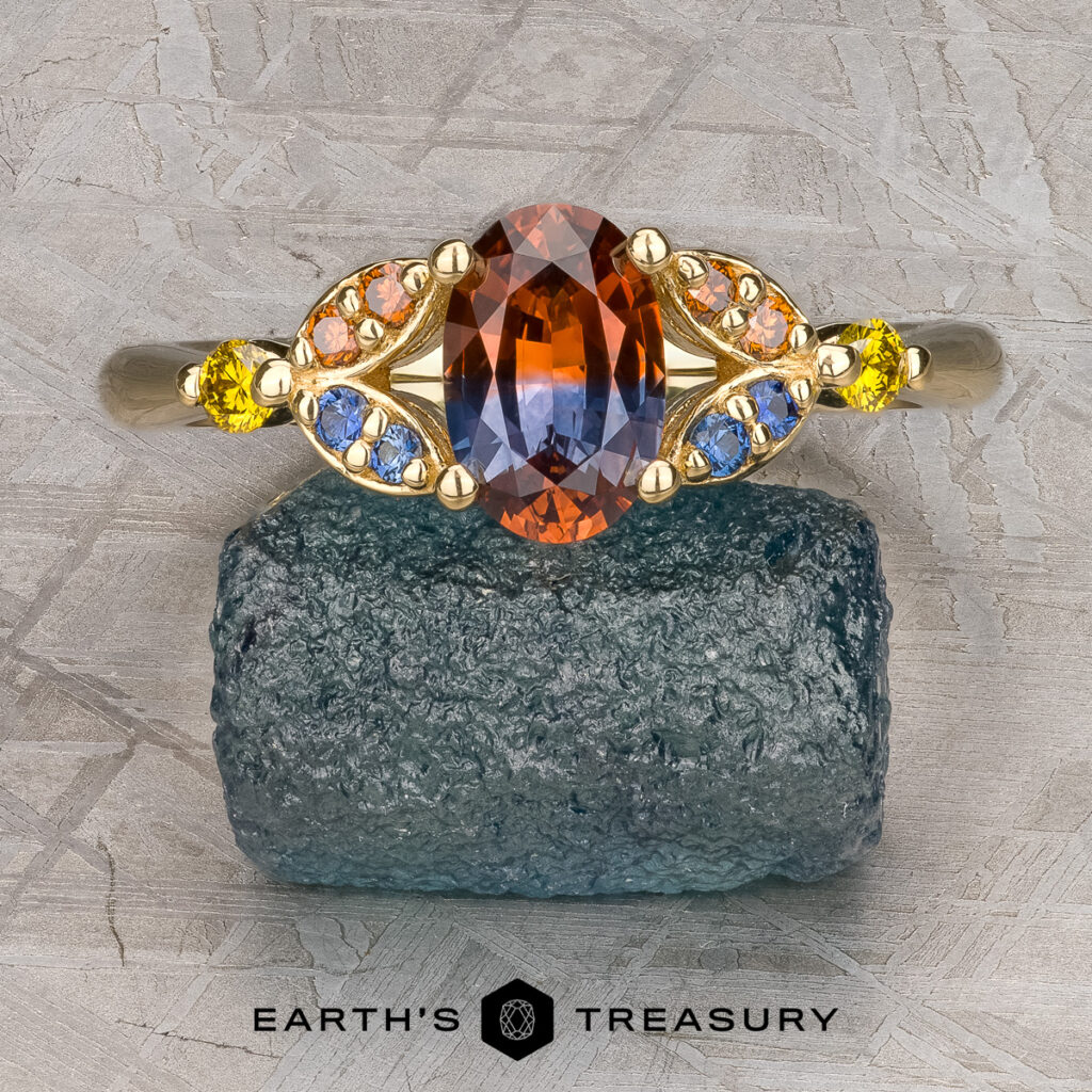 The Nabavi, Ali and Baran, Heather 10 custom gallery features a gold ring with an oval bi-color gemstone and yellow, orange, and blue accent stones displayed on a rough mineral above the "Earth's Treasury" logo.
