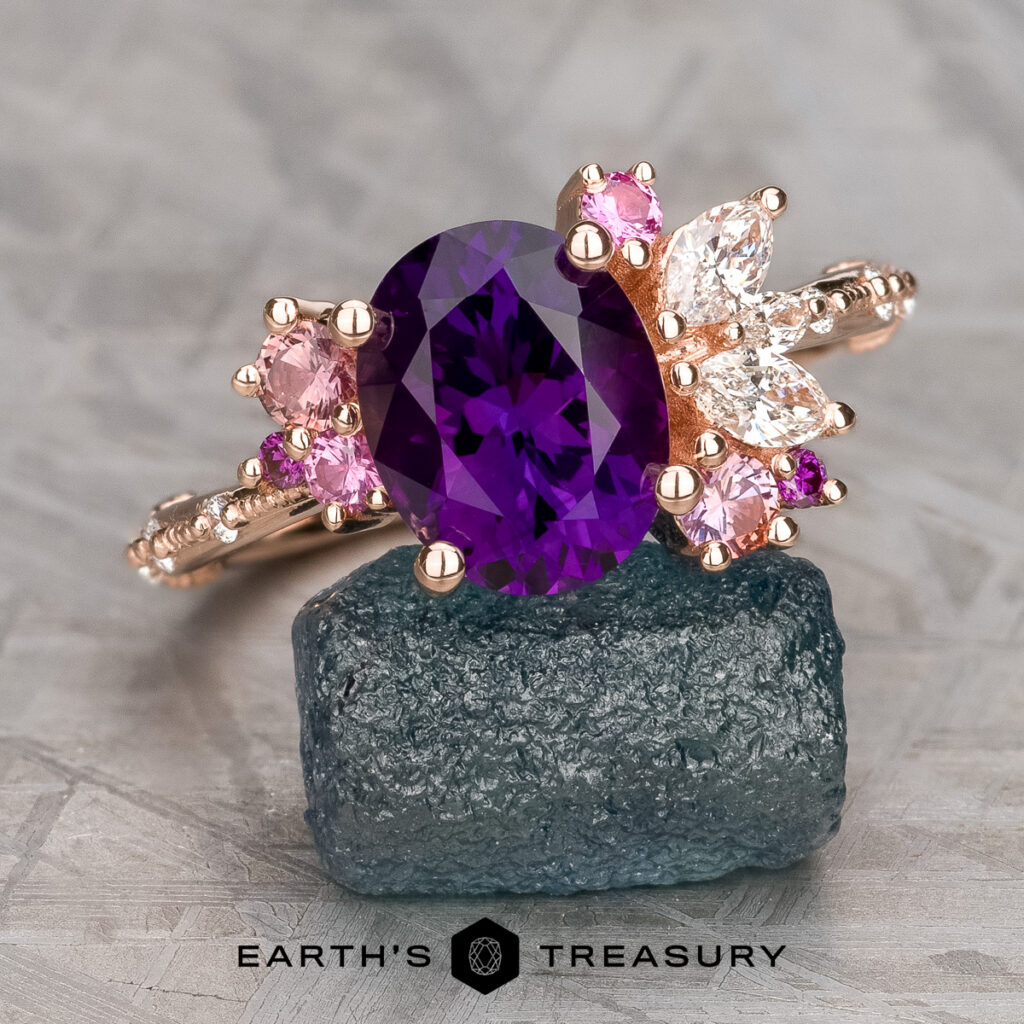 The Nabavi, Ali and Baran, Heather 13.5 gold ring features a large oval purple gemstone accented by pink and clear stones, displayed on a rough blue crystal with the Earth's Treasury logo below.