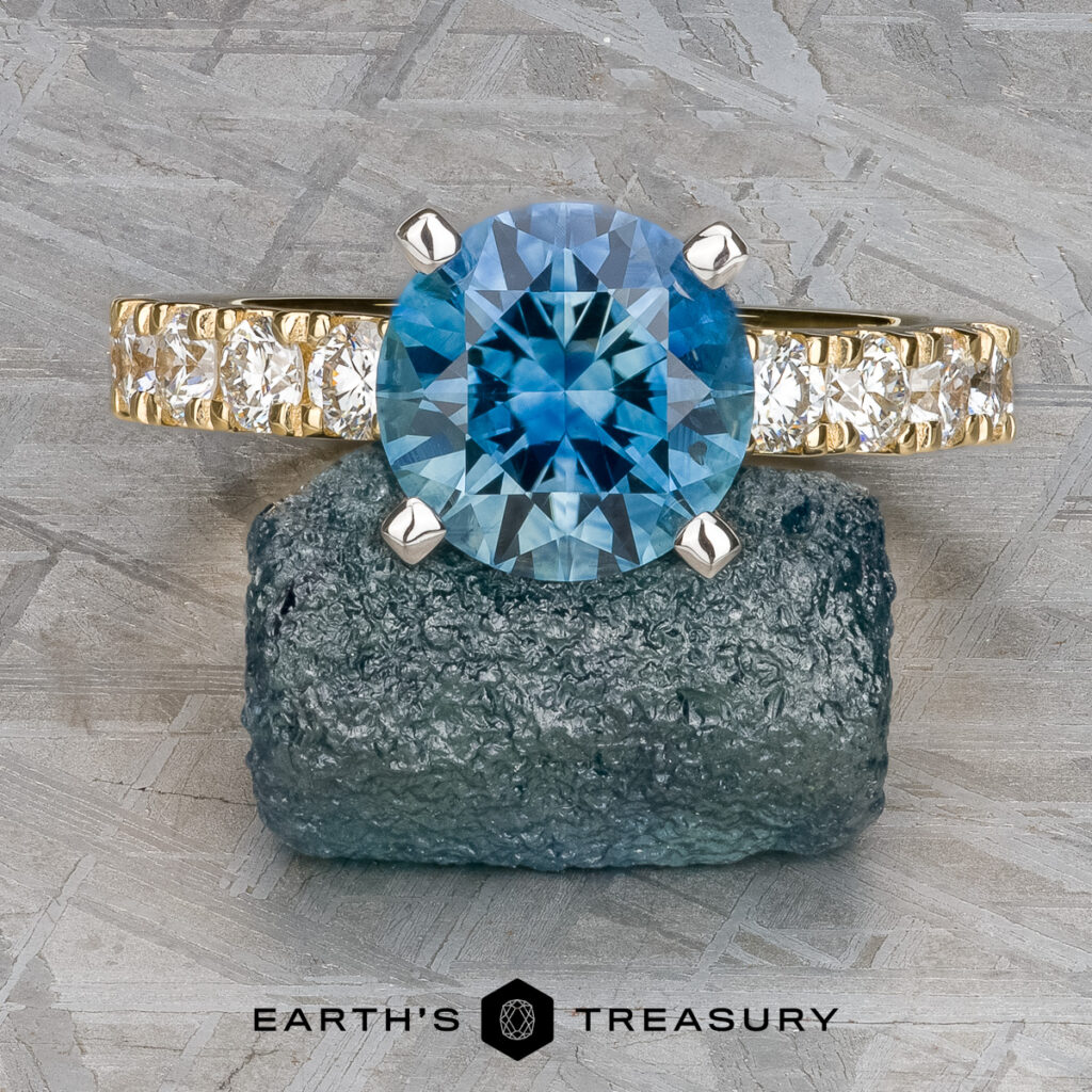 A gold ring with a large round blue gemstone and smaller white stones is displayed on a rough greenish rock, with the name "Thill, Thomas (Tom)" below.