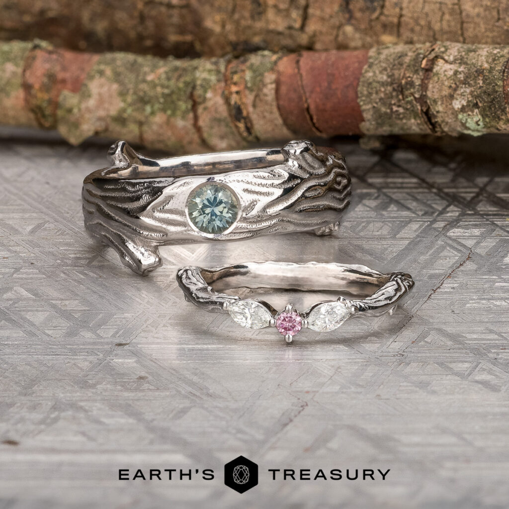 Two silver rings, “Stacked from 38 images. Method=B (R=11,S=7),” with organic designs rest on a patterned surface; one showcases a blue stone, the other a pink center stone flanked by two clear stones.
