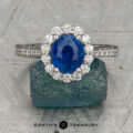 The "Josephine" ring in 18K white gold, featuring a 1.60-carat Ceylon sapphire with a prong-set halo of diamonds, is displayed on a blue-green gemstone against a patterned gray background with the "Earth's Treasury" logo below.