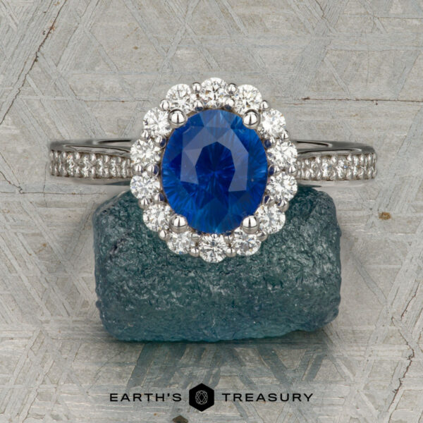 The "Josephine" ring in 18K white gold with 1.60-carat Ceylon sapphire The "Josephine" ring in 18K white gold, featuring a 1.60-carat Ceylon sapphire with a prong-set halo of diamonds, is displayed on a blue-green gemstone against a patterned gray background with the "Earth's Treasury" logo below.
