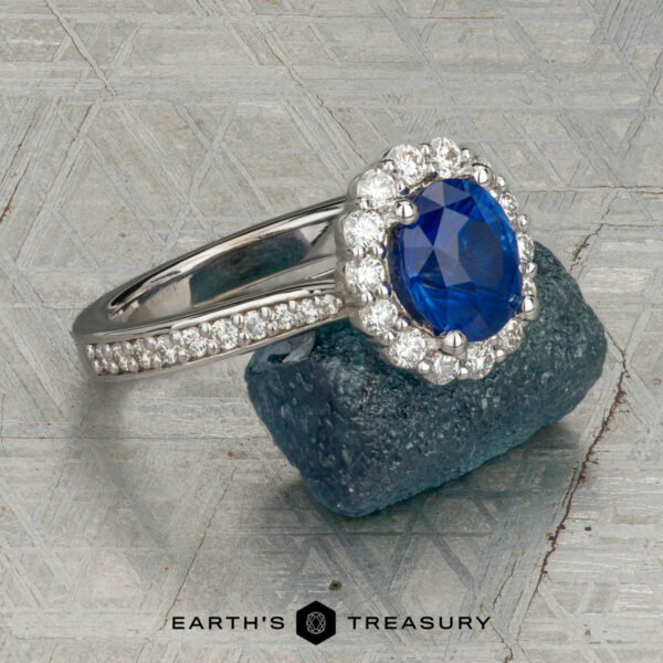 The "Josephine" ring in 18K white gold with 1.60-carat Ceylon sapphire The "Josephine" ring in 18K white gold, featuring a 1.60-carat Ceylon sapphire with a prong-set halo of diamonds, is displayed on a blue-green gemstone against a patterned gray background with the "Earth's Treasury" logo below.