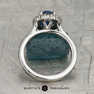 The "Josephine" ring in 18K white gold, featuring a 1.60-carat Ceylon sapphire with a prong-set halo of diamonds, is displayed on a blue-green gemstone against a patterned gray background with the "Earth's Treasury" logo below.