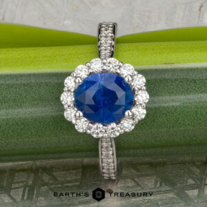 The "Josephine" ring in 18K white gold, featuring a 1.60-carat Ceylon sapphire with a prong-set halo of diamonds, is displayed on a blue-green gemstone against a patterned gray background with the "Earth's Treasury" logo below.