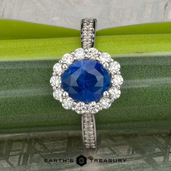 The "Josephine" ring in 18K white gold with 1.60-carat Ceylon sapphire The "Josephine" ring in 18K white gold, featuring a 1.60-carat Ceylon sapphire with a prong-set halo of diamonds, is displayed on a blue-green gemstone against a patterned gray background with the "Earth's Treasury" logo below.