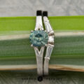The Long, Brant and Kucharczyk, Michelle 1 & 2 ring features white gold with a round blue-green gemstone, accented by baguette and round diamonds, shown against a green leaf backdrop.