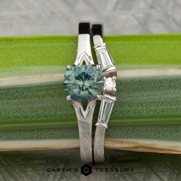 The "Cybele" ring in 18K white gold with 1.02-carat Montana sapphire, alongside the "Narelle" band in 18K white gold The Long, Brant and Kucharczyk, Michelle 1 & 2 ring features white gold with a round blue-green gemstone, accented by baguette and round diamonds, shown against a green leaf backdrop.