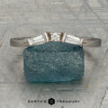 The "Narelle" band in 18K white gold, featuring a round center diamond and two tapered baguette diamonds, is displayed on a rough blue gemstone with the "Earth's Treasury" logo below.