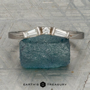 The "Narelle" band in 18K white gold, featuring a round center diamond and two tapered baguette diamonds, is displayed on a rough blue gemstone with the "Earth's Treasury" logo below.