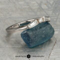 The "Narelle" band in 18K white gold, featuring a round center diamond and two tapered baguette diamonds, is displayed on a rough blue gemstone with the "Earth's Treasury" logo below.