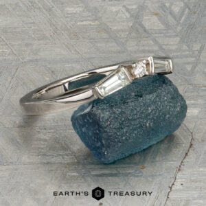 The "Narelle" band in 18K white gold, featuring a round center diamond and two tapered baguette diamonds, is displayed on a rough blue gemstone with the "Earth's Treasury" logo below.