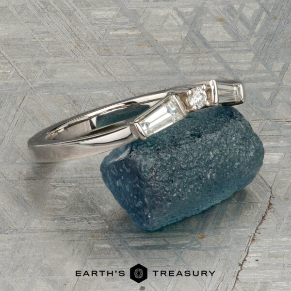 The "Narelle" band in 18K white gold The "Narelle" band in 18K white gold, featuring a round center diamond and two tapered baguette diamonds, is displayed on a rough blue gemstone with the "Earth's Treasury" logo below.