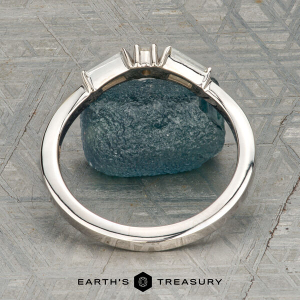 The "Narelle" band in 18K white gold The "Narelle" band in 18K white gold, featuring a round center diamond and two tapered baguette diamonds, is displayed on a rough blue gemstone with the "Earth's Treasury" logo below.