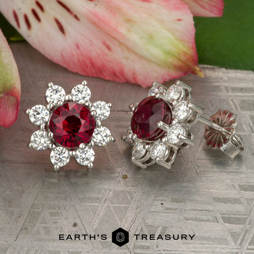 A pair of Lao, Vicky flower-shaped earrings with red center stones and clear gemstones, displayed on a textured surface with a flower and green leaf in the background.
