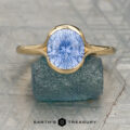 The Hansen, Cameron ring features a round, starburst-cut light blue gemstone set in gold, displayed upright on a rough blue stone with the Earth's Treasury logo below.