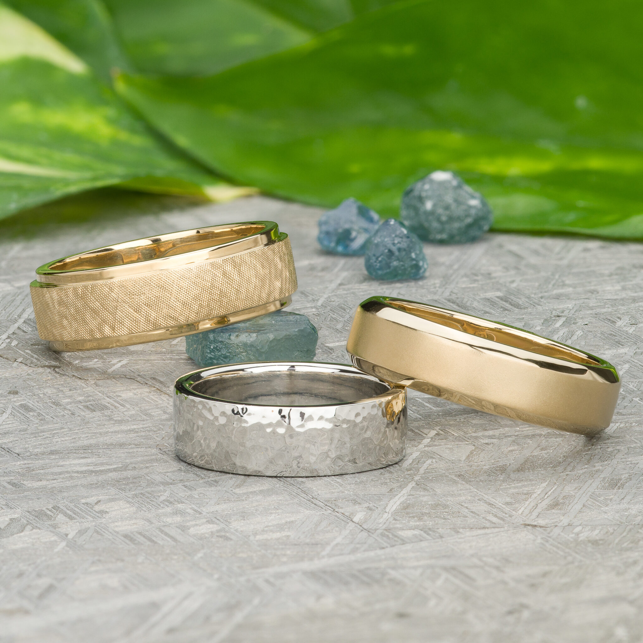 The product "Stacked from 74 images. Method=B (R=7,S=2)" features three wedding bands—matte gold, hammered silver, and polished gold—on a textured surface with green leaves and blue stones in the background.