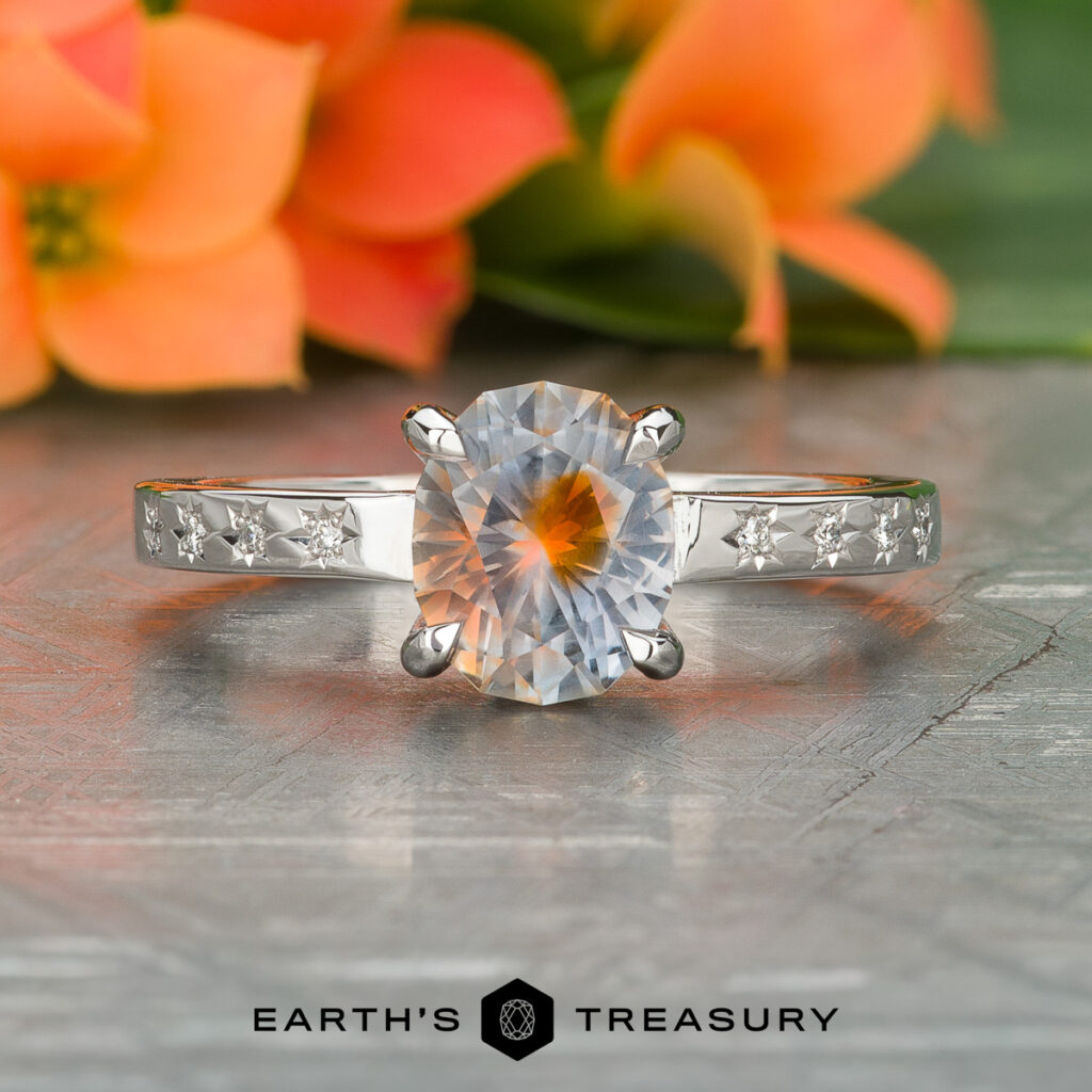 The Feldman, Gabriella [custom library] is a silver ring featuring an oval-cut gemstone and star engravings on the band, shown on a textured surface with orange flowers in the background.