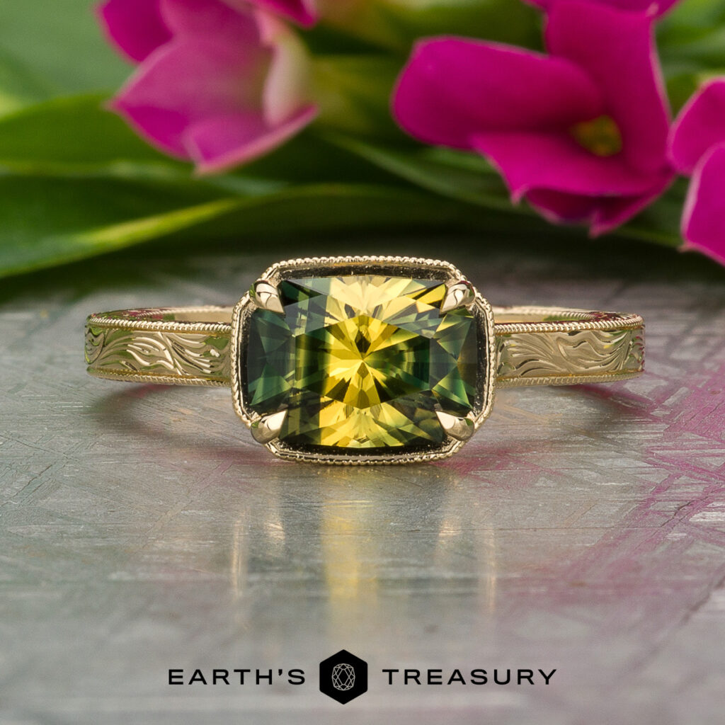 The Storrer, Carly and Meek, Hunter [Custom Gallery] ring features a square-cut yellow-green gemstone on an engraved gold band, displayed on a reflective surface with pink flowers in the background.