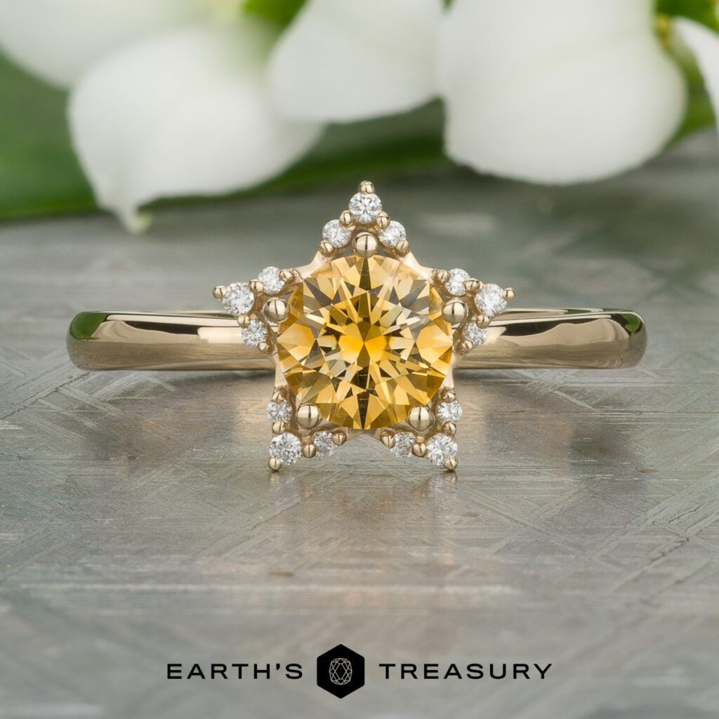 A custom gold ring from Chau, Austin and Castillo, Aida showcases a yellow star-shaped gemstone surrounded by small diamonds, set on a patterned surface with white flowers in the background. "EARTH'S TREASURY" logo appears below.