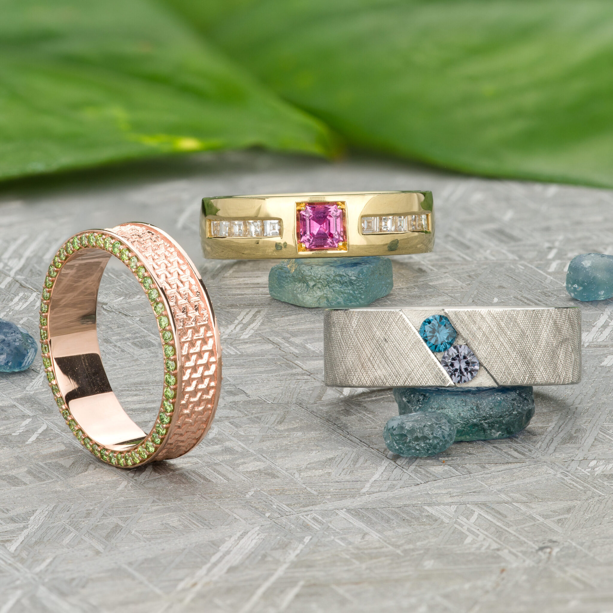 Three "Stacked from 87 images. Method=B (R=7,S=2)" rings on a textured surface: a rose gold band with green gems, a yellow gold ring with pink center stone, and a silver ring with blue and white gems among green leaves and blue stones.