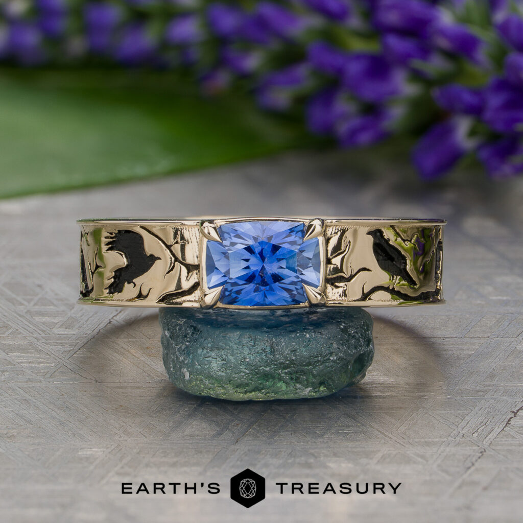 The Poirier, Bailey [custom gallery] gold ring with engraved designs features a square blue gemstone at its center, displayed on a green stone with purple flowers in the background and the Earth’s Treasury logo below.