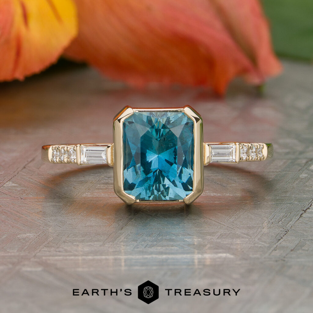 A Heckman, Christine gold ring featuring an emerald-cut blue gemstone at the center, accented by baguette and round diamonds, displayed on a patterned surface with floral decor in the background.
