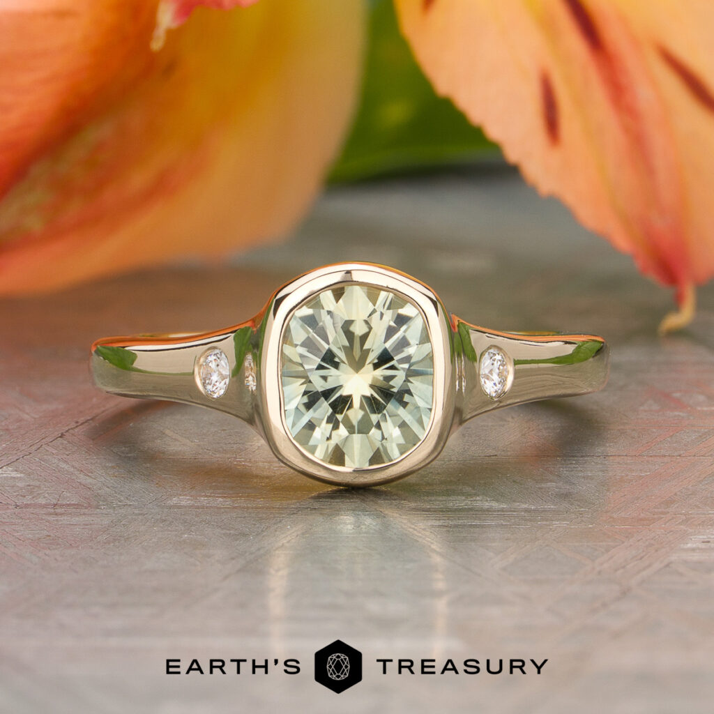 The TeeGarden Kaelib [custom library] gold ring features a large cushion-cut gemstone at its center with two small round diamonds on each side, displayed on a patterned surface with flowers in the background.