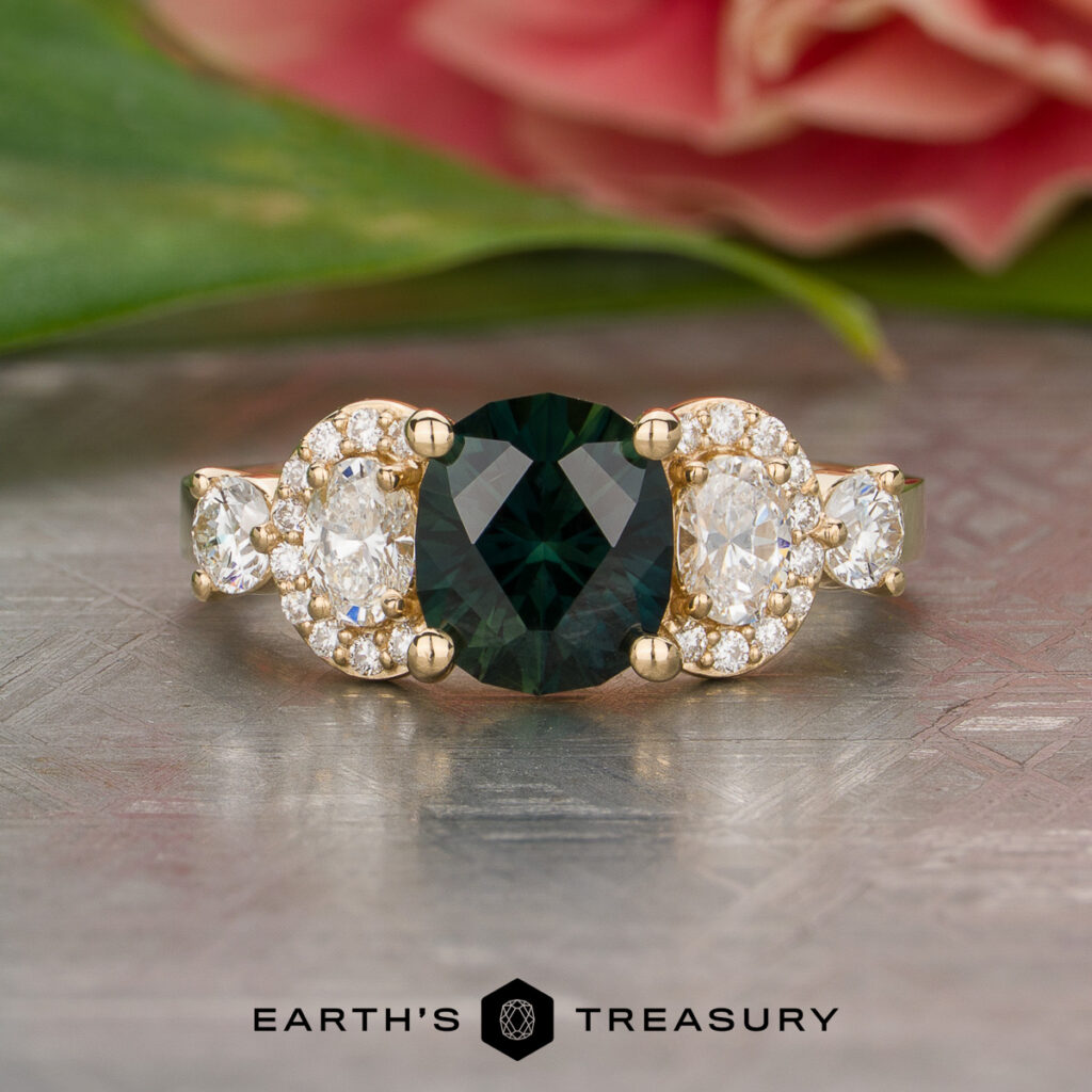 The Abukar Rahma [custom gallery] features a gold ring with an oval dark green center stone, accentuated by oval and round clear diamonds, showcased on a textured background with a flower nearby.