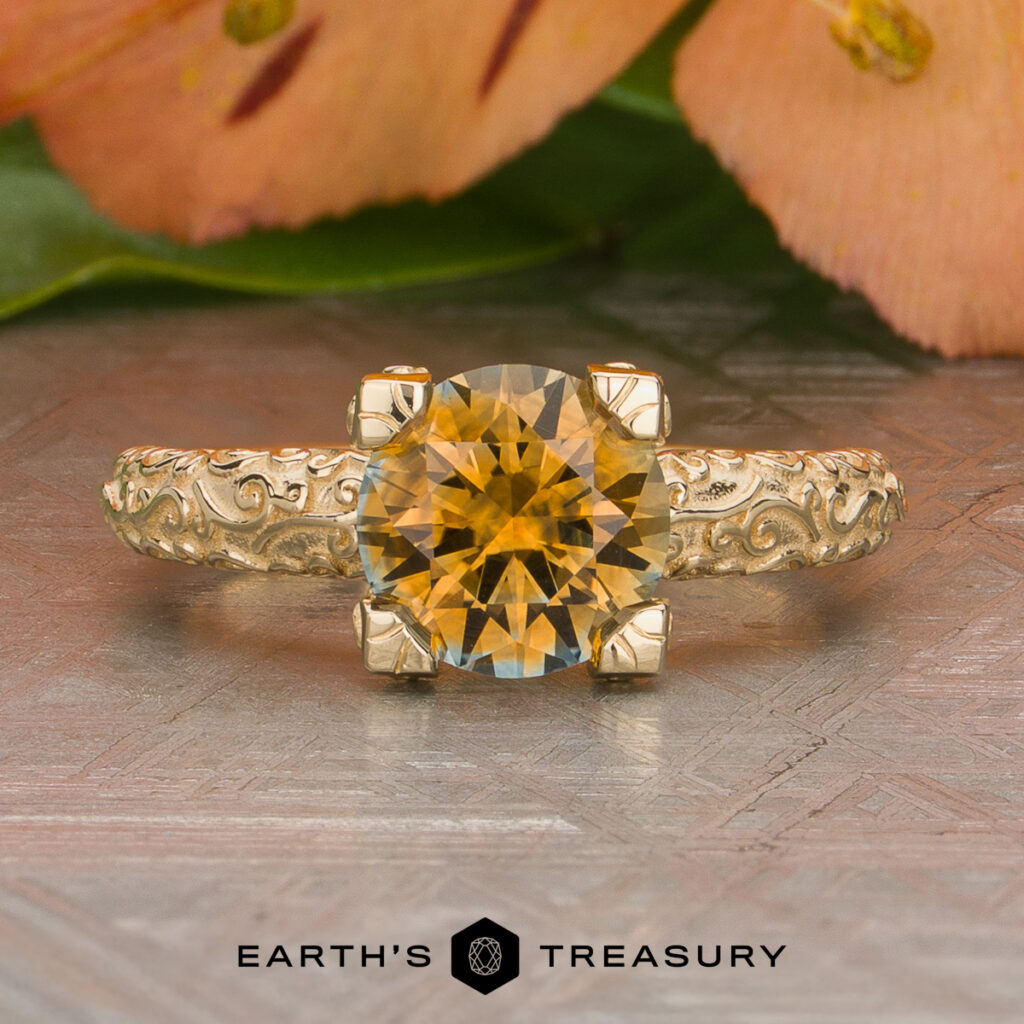 The Del Rio Victor ring features a round yellow gemstone in a prong setting on an ornate gold band, with soft pink flowers and green leaves in the background. The "Earth's Treasury" logo is displayed at the bottom.