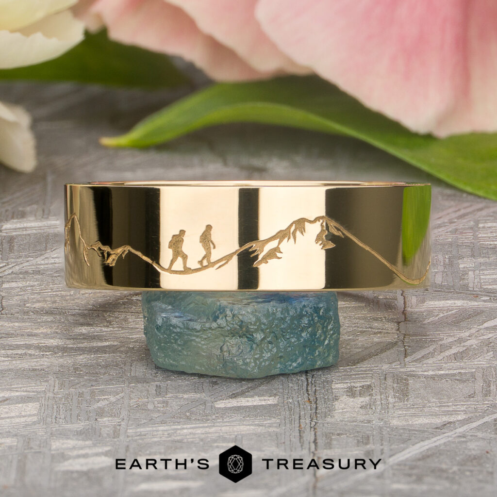 An Updike, Thomas gold ring engraved with a mountain range and two hikers, displayed on a rough gemstone with flowers in the background. The "Earth's Treasury" logo is shown below.