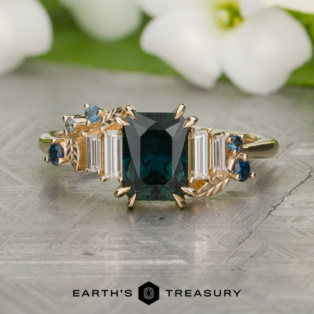 Custom gallery by Clarke, Liam and Burns, Sarah: Gold ring featuring a rectangular dark green gemstone center with baguette and round blue stones, displayed on a patterned surface with "Earth's Treasury" below.